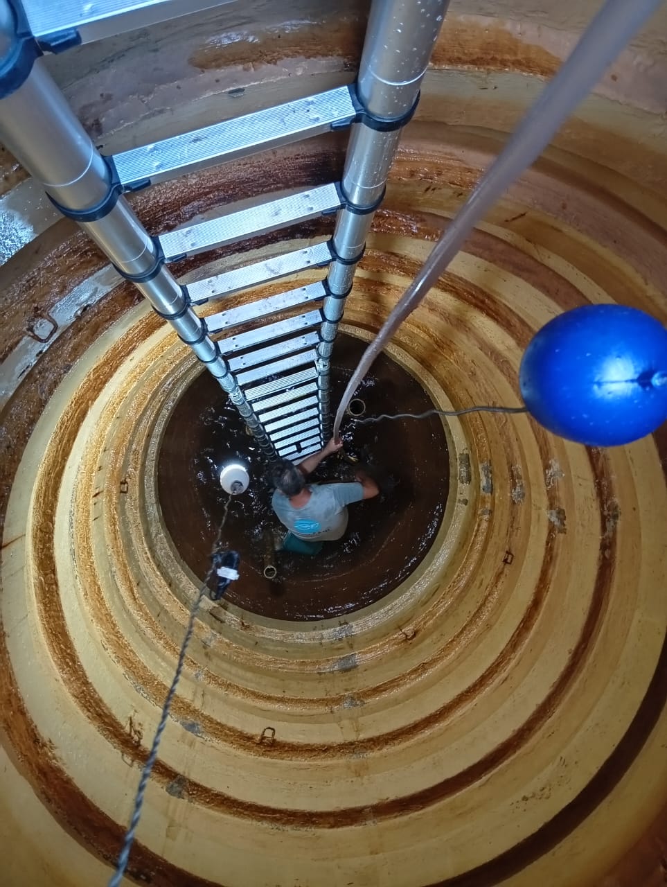 homem dentro de um poço/cisterna circular visto de cima, com escada de alumínio e boia azul na água